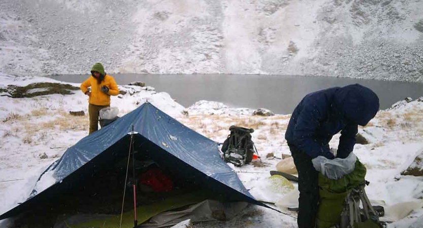 Two people stand near a tarp shelter that is resting beside a body of water. I light dusting of snow is covering the landscape.